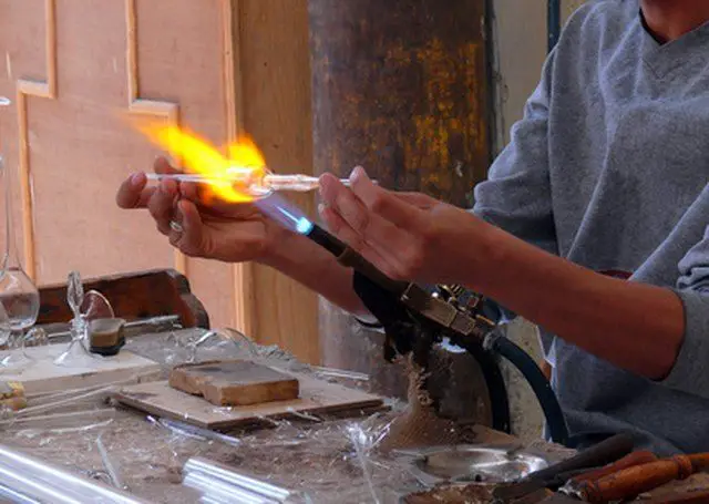 a person using a small torch to melt broken pieces of glass at a table in their garage.