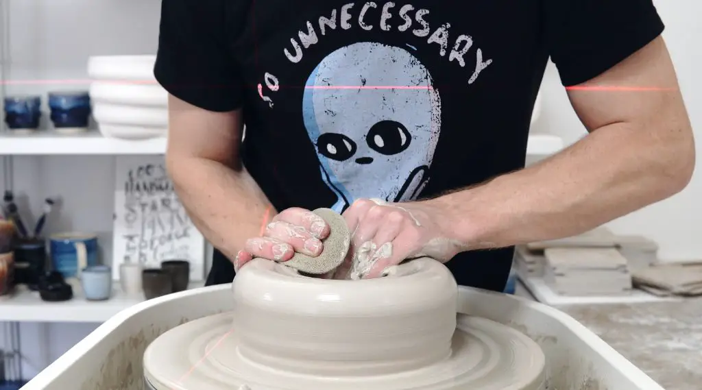 a potter wearing flexible yoga pants and a t-shirt while throwing a clay pot on the wheel