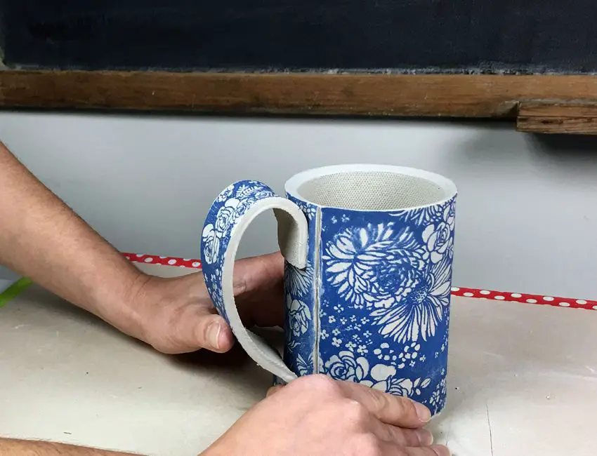 an artist carefully sketching out a design on a ceramic mug with an underglaze pencil