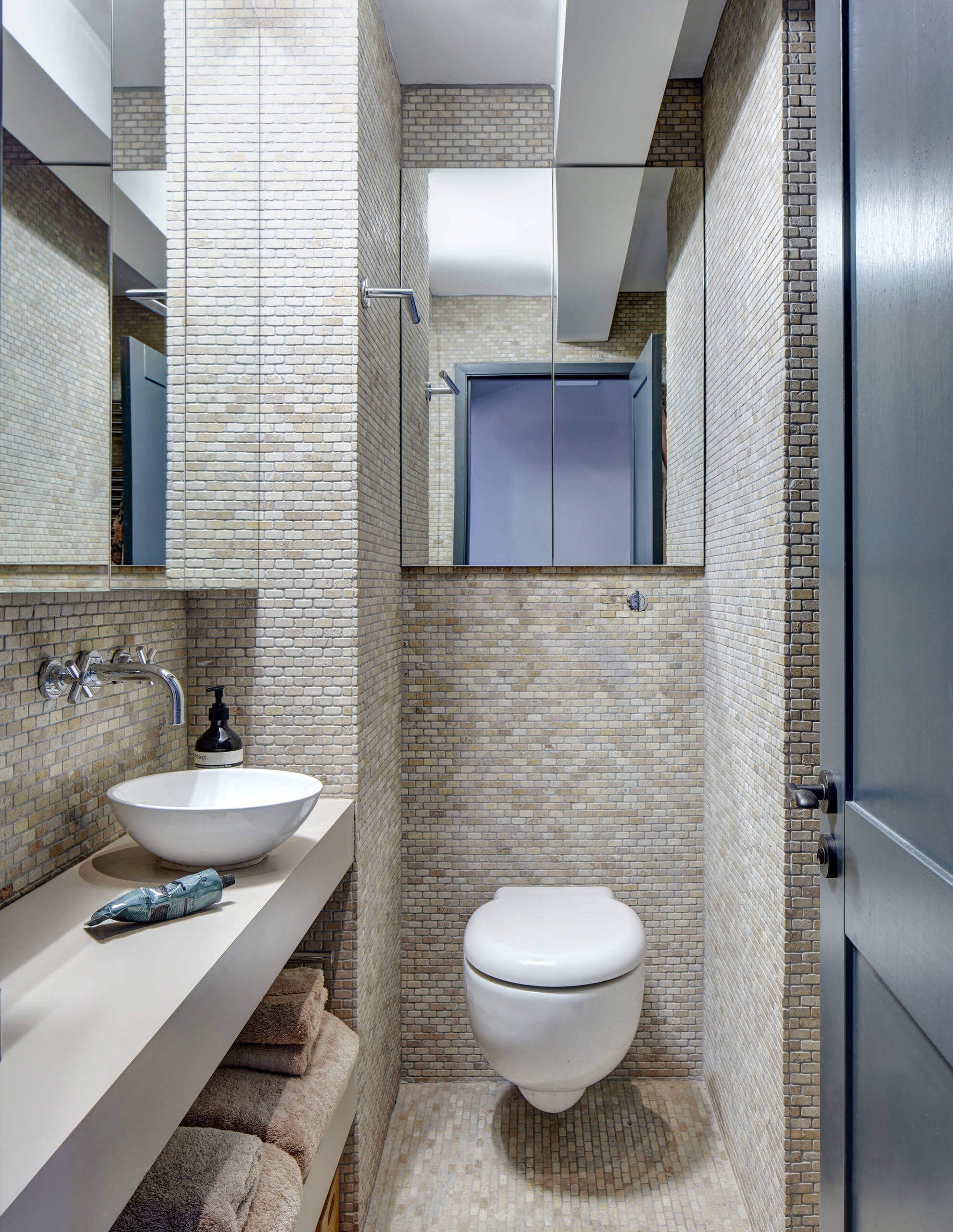 #10. Transitional beige tile bathroom remodel with a vessel sink, open cabinets and a wall-mount toilet